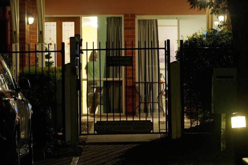 The inside of a brick aged care home is seen through windows and a tall fence illuminated at night.