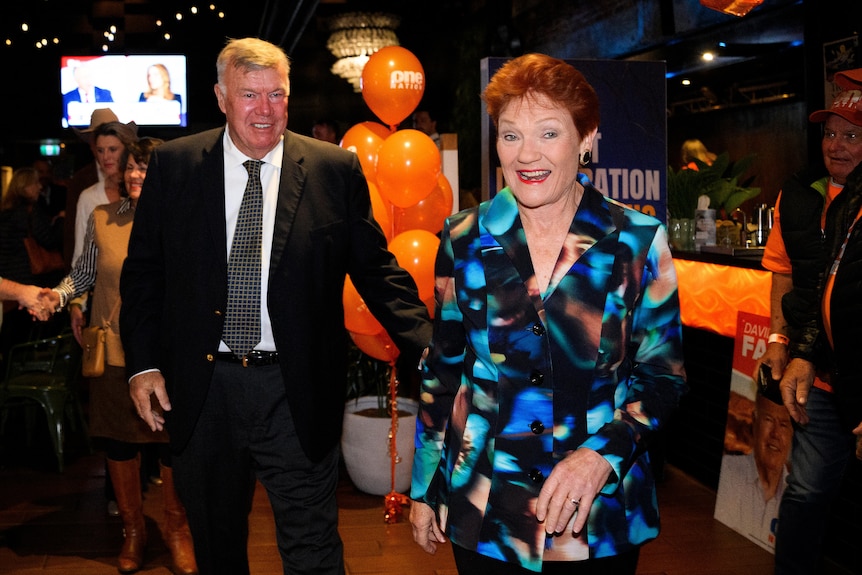 Woman in colourful dress leads man in a suit past orange balloons at a political victory party.