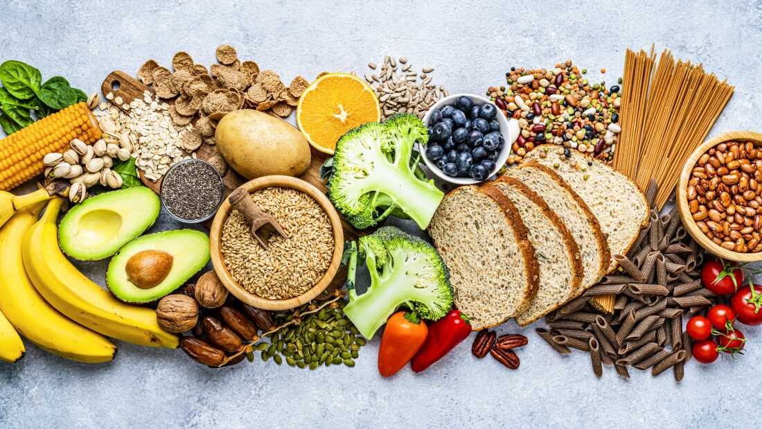 Overhead view of a large group of food with high content of dietary fiber arranged side by side. The composition includes blueberries, oranges, avocado, wholegrain bread, potatoes, oats, corn, beans, greens, broccoli, mixed nuts, and bananas, among others.