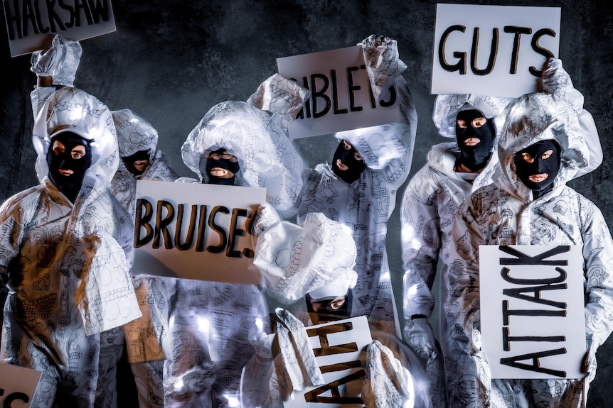 TISM's seven members wear face masks and brandish signs reading BRUISE, ATTACK, GUTS and more