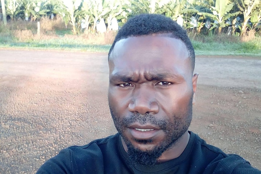 A selfie image of a man wearing a black top on a tropical farm.