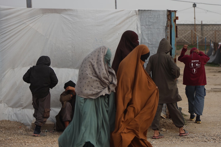 A group of three women wearing burqas walk along a camp in Syria.