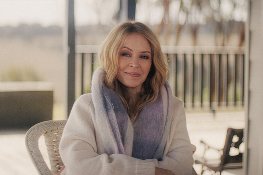 pop star Kylie Minogue sits in a dressing gown on a deck chair smiling pensively. 