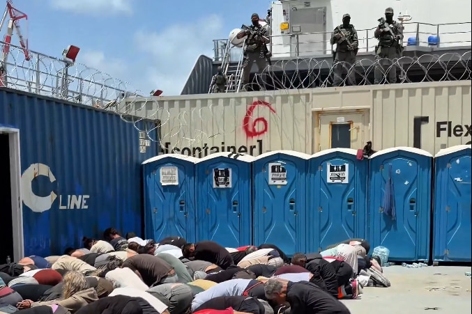 people kneeling on the ground as armed soldiers watch over