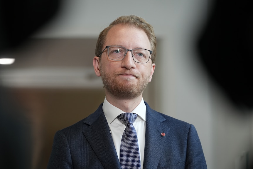 James Paterson stands wearing a suit looking serious.
