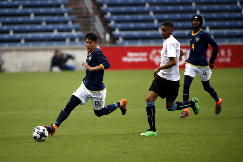 Three male players on the field, one controlling the ball with his teammate following, and a member for the opposing teams is coming up behind him to take the ball.