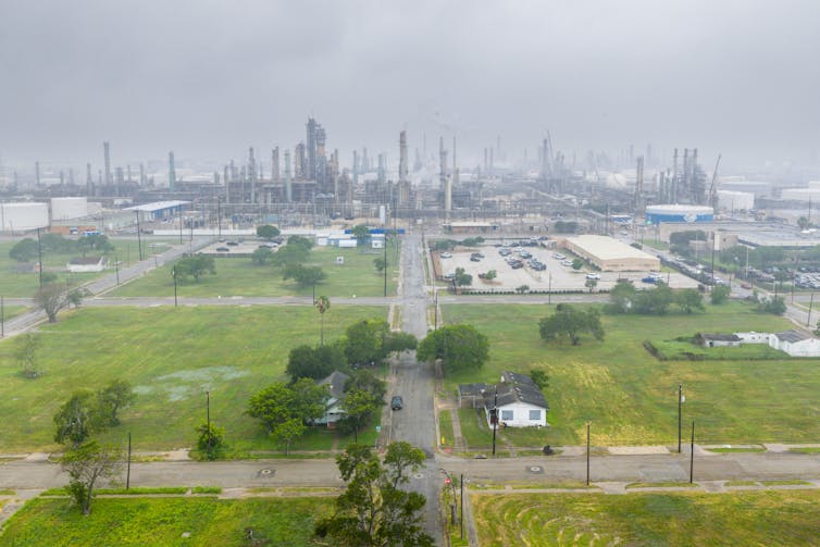 A neighborhood with grassy lawns and trees is in the foreground, with a massive industrial complex in the background.