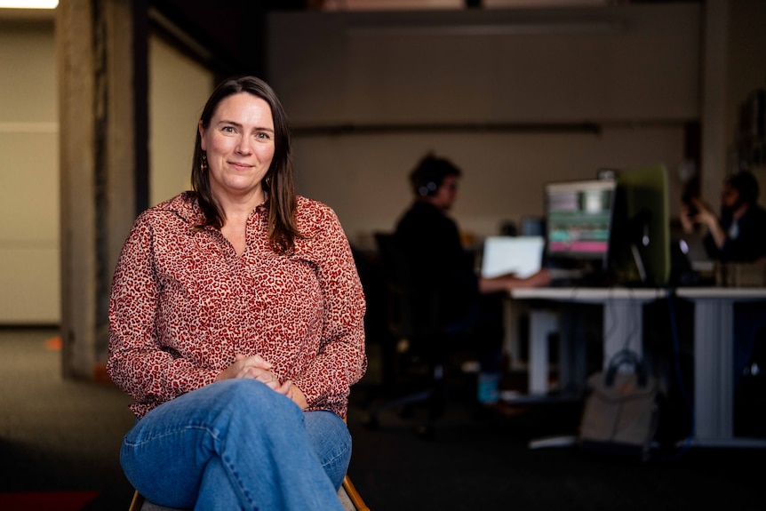 A woman smiles for a photo, sitting in a room with computers behind her