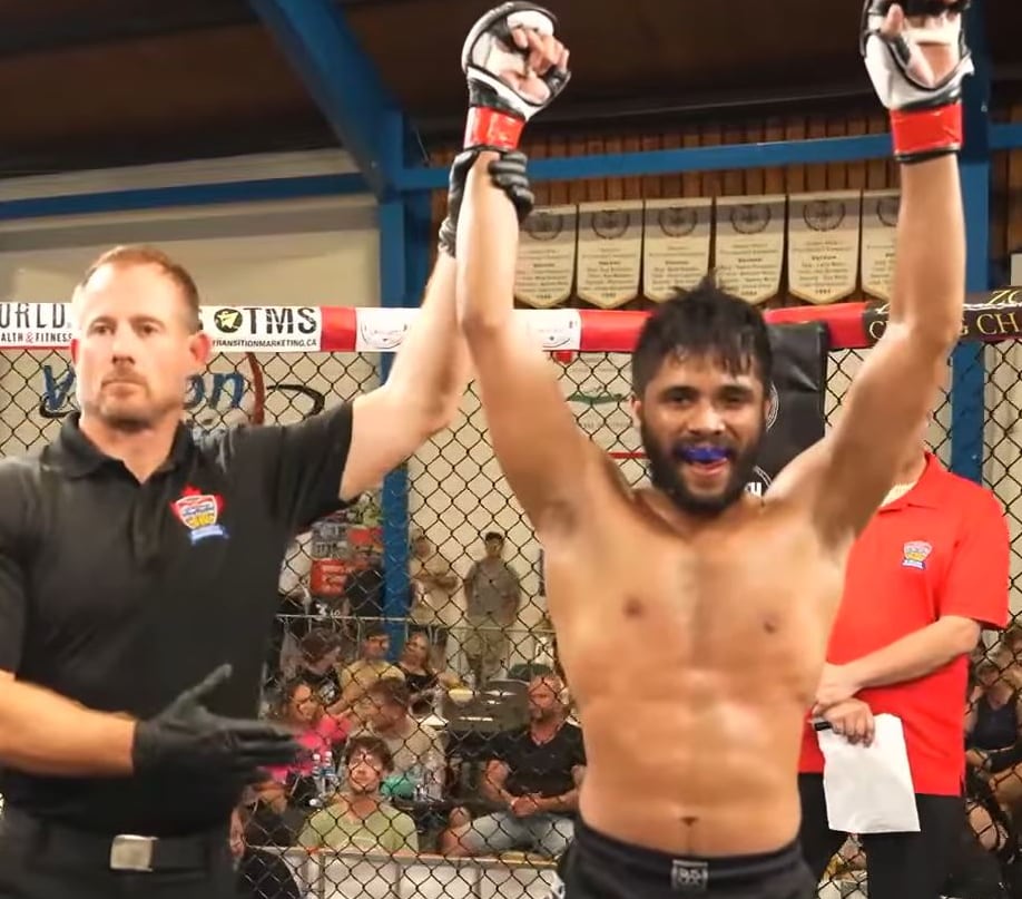 A martial arts referee holds up the arm of a shirtless man standing in a fighting ring.