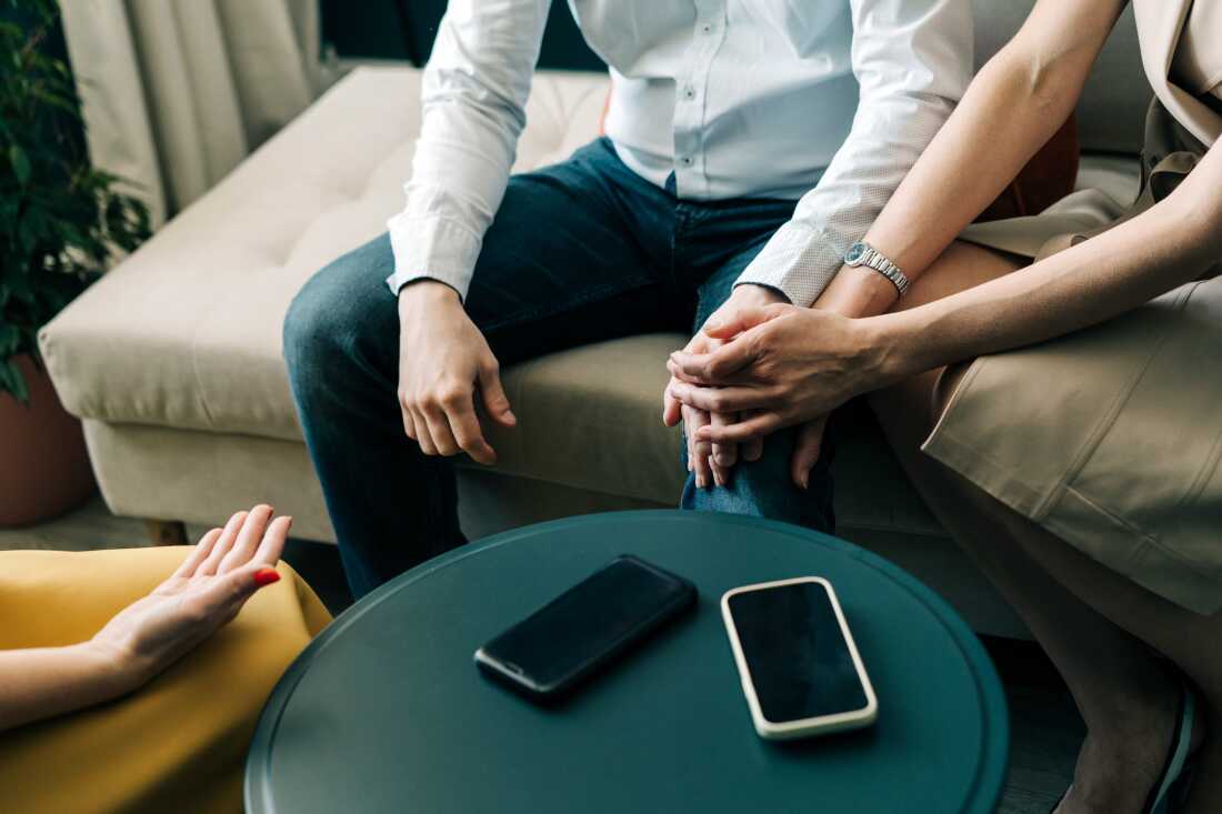 This photo shows a couple — man and woman — sitting next to one another and holding hands. Seated across from them is a female mental health therapist. Between the two parties is a round coffee table with two smartphones on it. The three people are photographed from about the shoulders down, so their faces don't show.