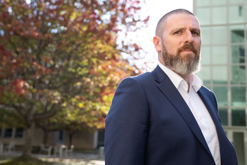 A man with short hair and a greying beard looks to the left of camera. He wears a white collared shirt and navy jacket 