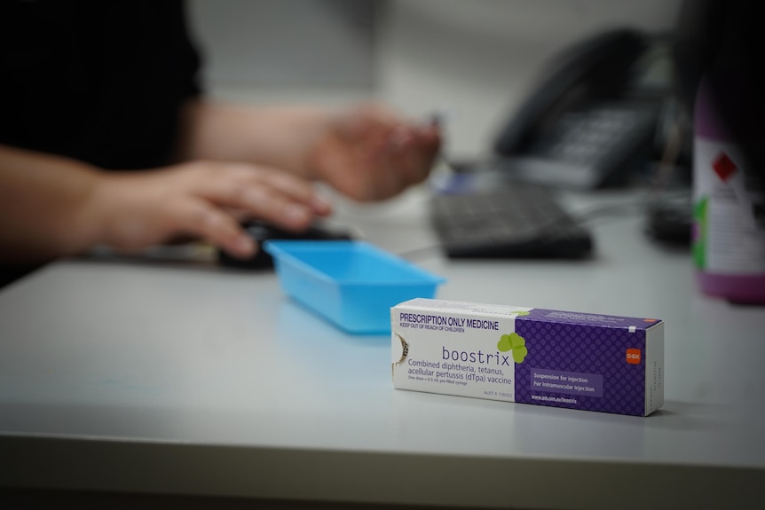 A nurse preparing a boostrix vaccination, the vaccine's white and purple box is visible in the foreground.