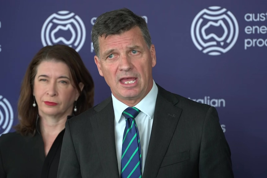 Angus wearing a suit and tie speaks with a woman in the background and a blue banner for Australian Energy Producers