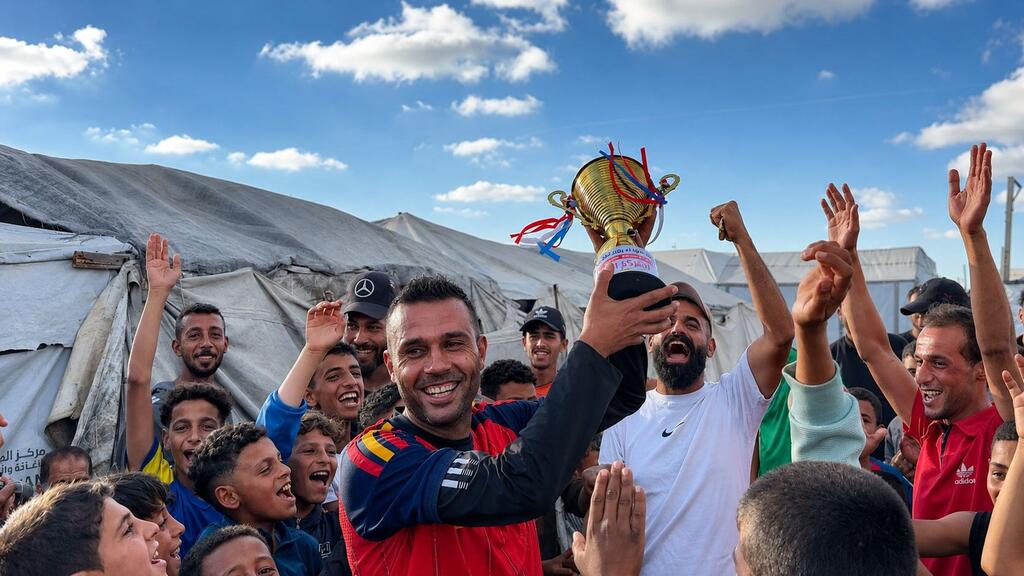 A Palestinian football player lifts a soccer trophy in a refugee camp in Gaza, surrounded by celebrating teammates and children.