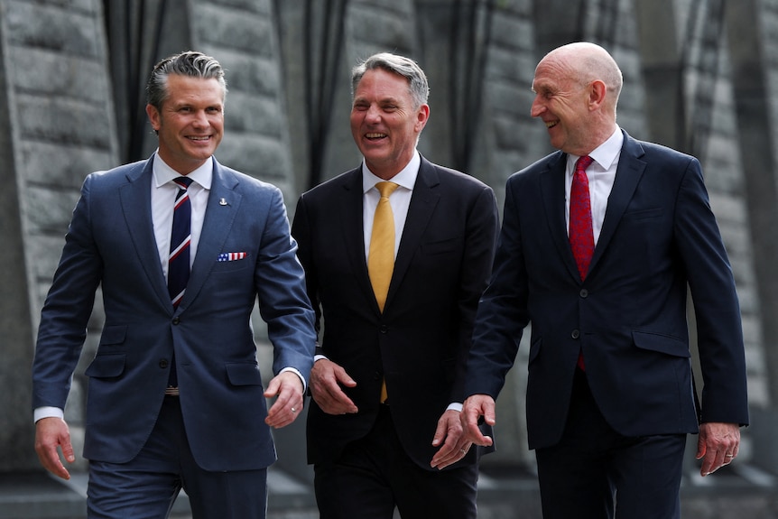 Three men in suits and ties walk together outside with happy expressions.