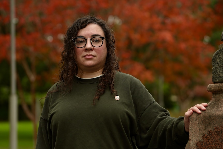 A woman with a serious expression brown hair, glasses and a dark green jumper in a park