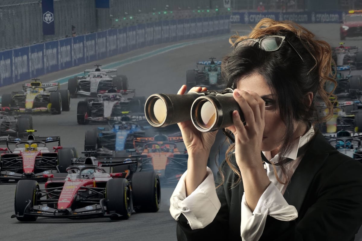 A woman looks through a pair of binoculars to the background of F1 cars