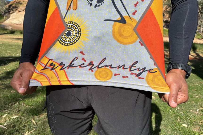 Two black sleeved arms hold out the bottom of a shirt which show the word Irrkerlantye on the bottom.