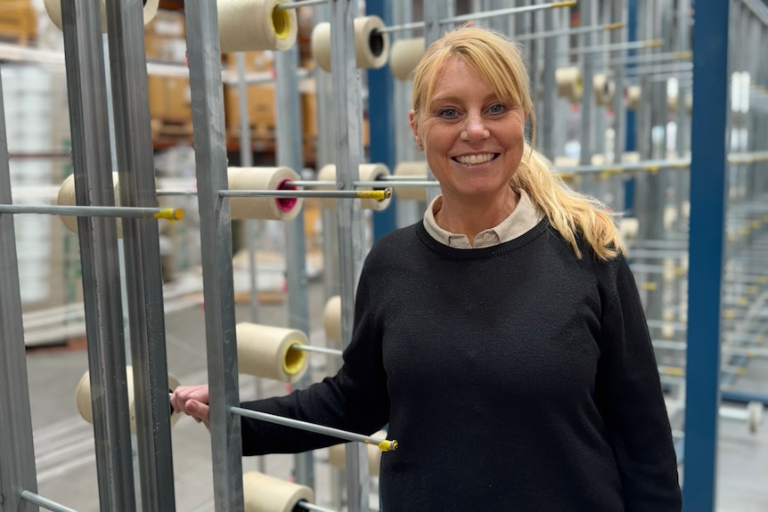 A young blonde woman grins standing next to metal spikes with large spools of thread