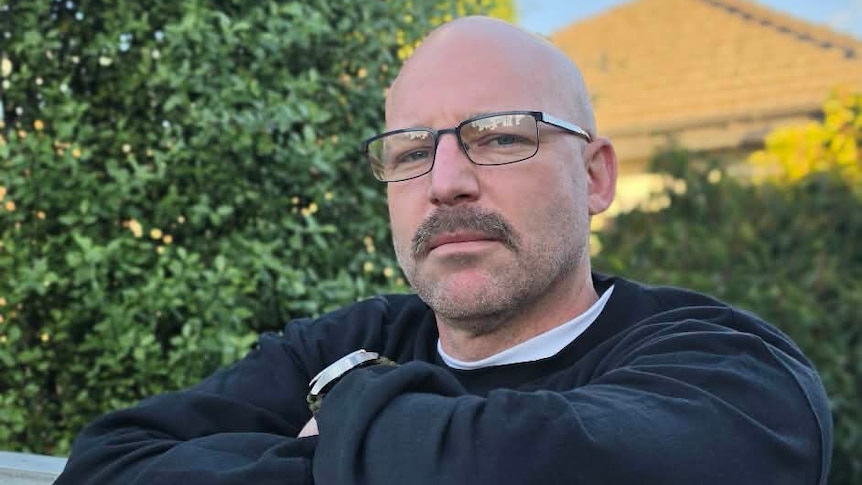 A bald man wearing glasses and a navy top rests his folded arms on a white wooden fence near trees and stares at the camera.