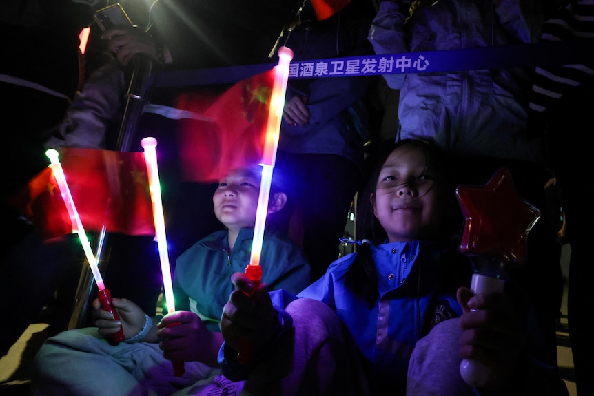 Chinese children waving illuminated flag wands while sitting, and their faces lit up in a dim blue light.