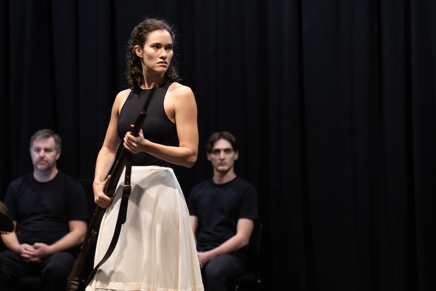 Nina Korbe holds a rifle at hip height with a serious expression. She wears a black top and long white skirt.
