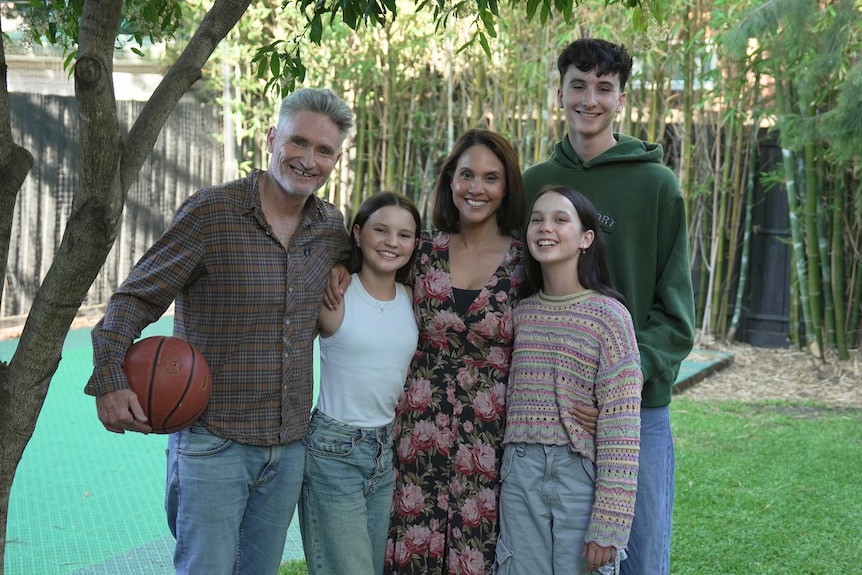 Two adults and three teenagers stand embracing in a suburban backyard.