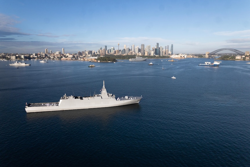 A grey frigate is seen in the foreground, sailing in Sydney Harbour, with the bridge to the right of the picture.