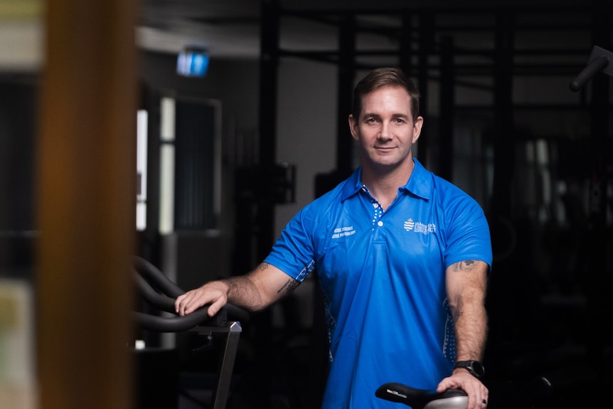 A man wearing a blue polo shirt smiling in front of gym equipment.