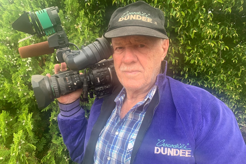 Man stands in front of bush wearing hat and jumper and holding camera over shoulder