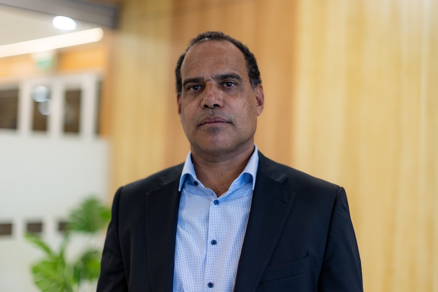 A man with short dark curly hair wearing a navy blue jacket and a light blue shirt.