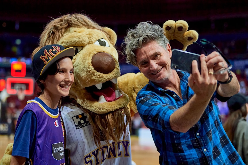 Dave Hughes poses with his teenaged son and a mascot on a basketball court