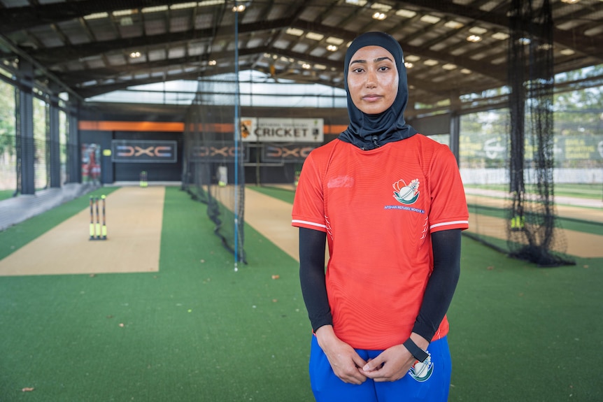 An Afghan woman, tanned, black hijab, red polo shirt, black sleves, standing in front of three sheltered cricket pitches.