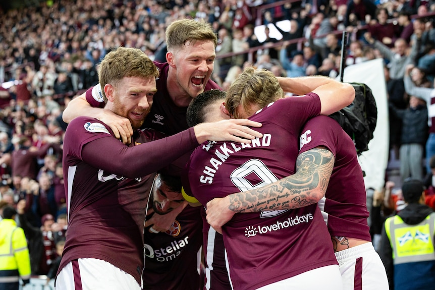 Four soccer players embrace after a goal, in front of cheering fans