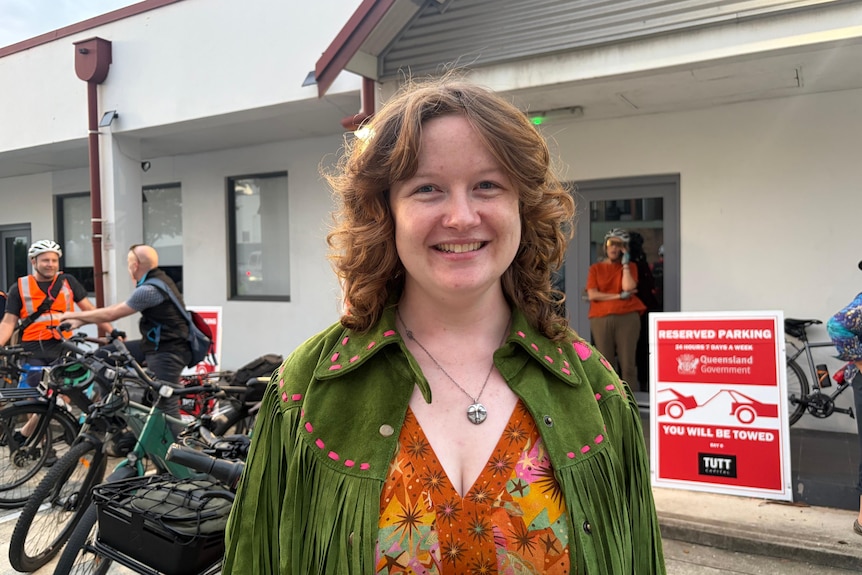 A woman wearing bright orange and a green jacket smiles with bikes parked in the background.