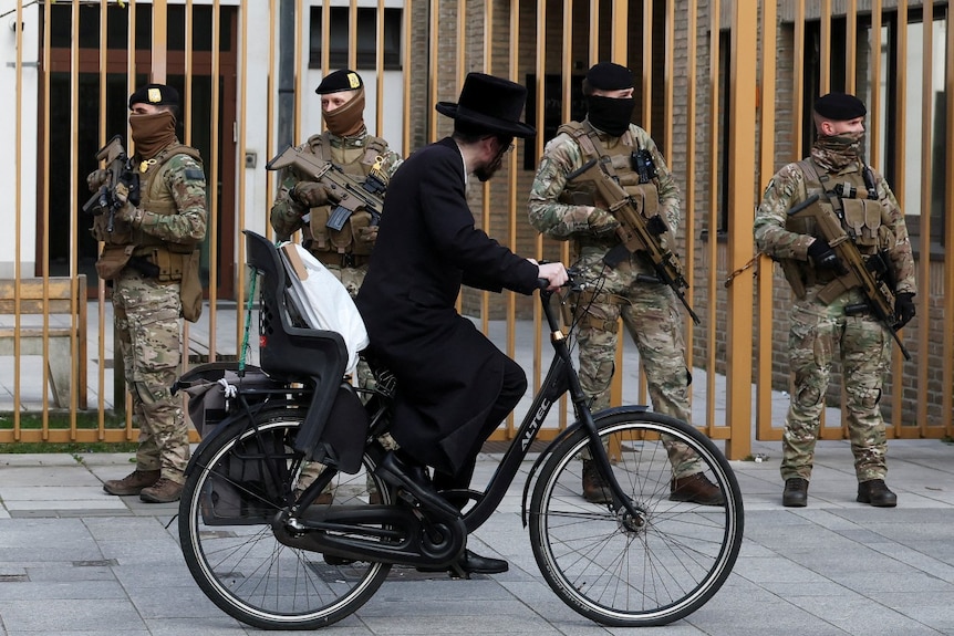 A man wearing religious garb cycles past several soldiers.