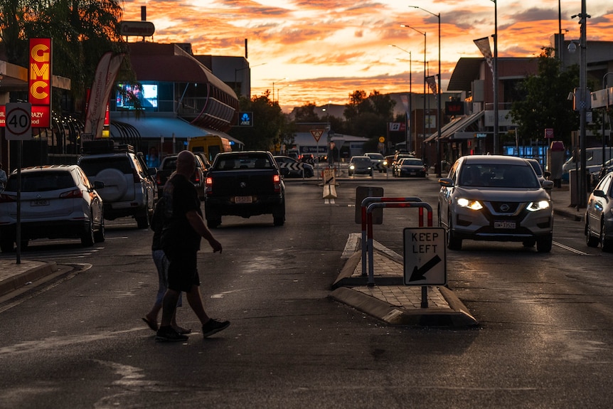 People and cars on a busy street in the Alice Springs CBD as the sun sets on the horizon.