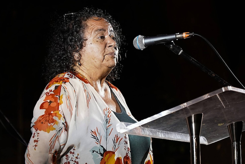A woman with dark curly hair speaks at a lectern at an evening ceremony.