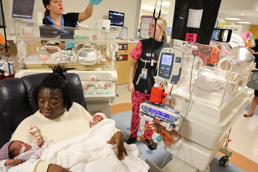 A woman cradling two newborns with medical staff and incubators behind her