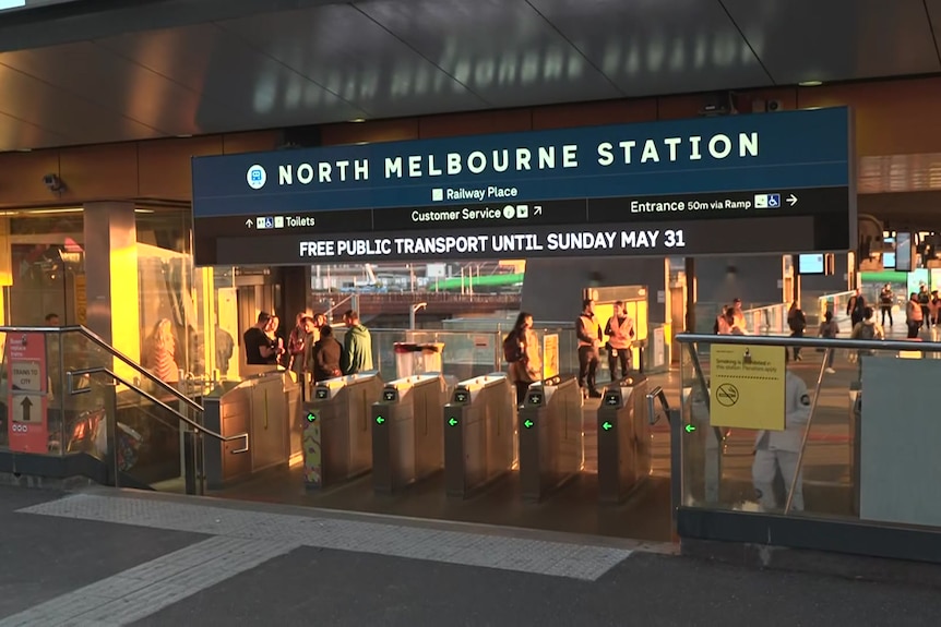 People stand behind entry gates to a railway station under a sign that says "North Melbourne Station" in the late afternoon sun.