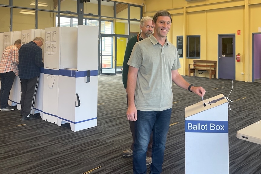 A man wearing a green shirt and jeans smiles, placing paper into a ballot box.