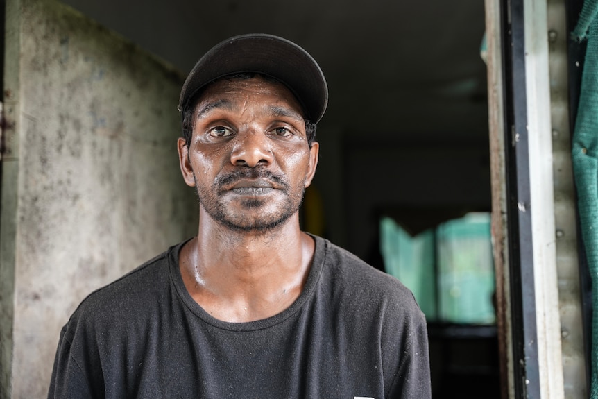 An Aboriginal man wearing a peak cap and black t-shirt looking at camera