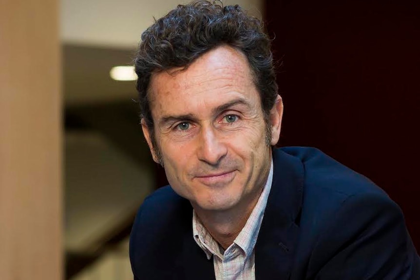 A man with dark wavy hair in a blue blazer and white collared shirt looks at the camera with a closed lipped smile.