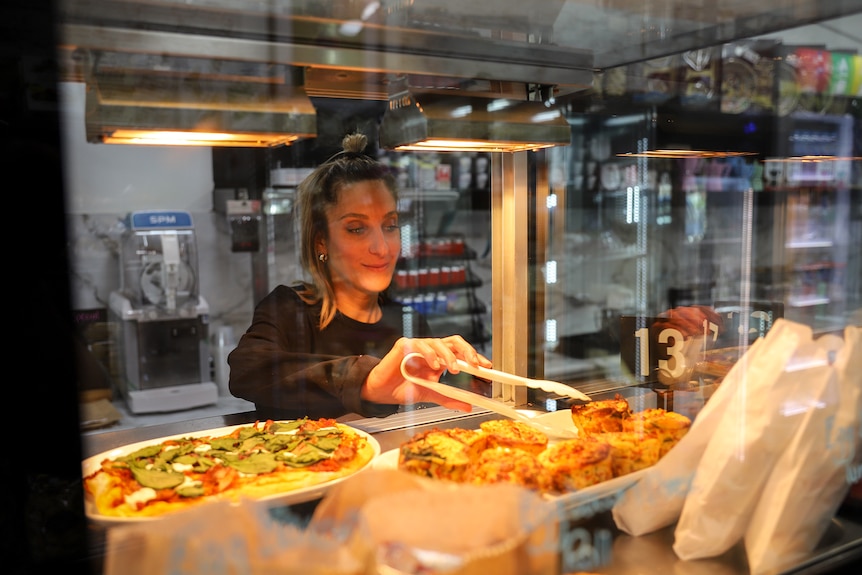 A woman stands behind a glass cabinet and holds a pair of tongs using them to pick up a baked good.