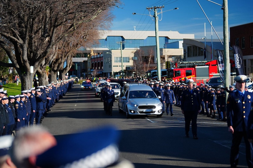 View of a funeral procession, with police officers lining a street as a hearse passes by.