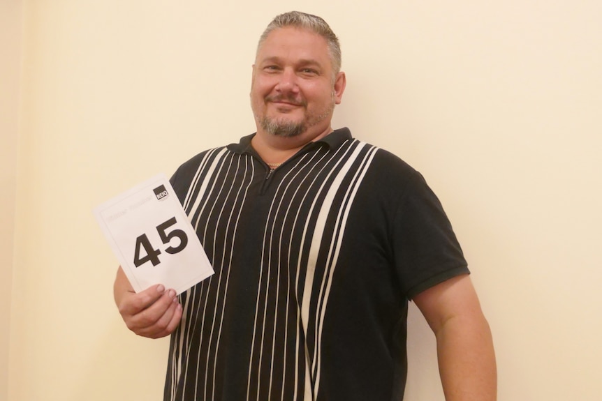 Large man in stripe polo shirt in front of blank wall, holding bid 45 card, smiling