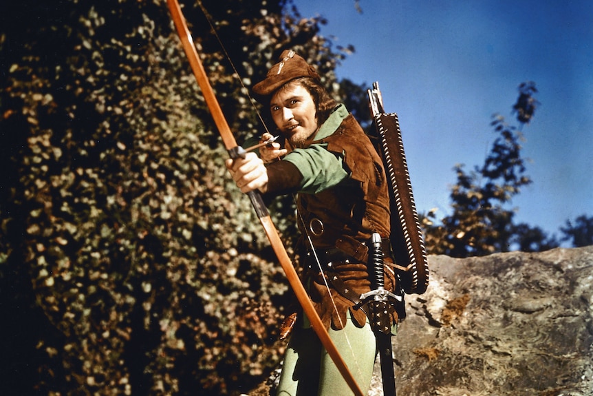 Errol Flynn poses in costume in Robin Hood, taking aim with a bow-and-arrow.