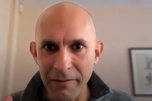 Close-up shot of Anil looking directly at the camera. Framed pictures in the background.