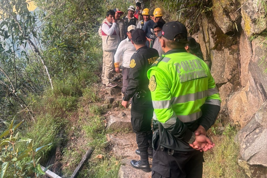Peruvian police in hi-viz or black clothing and yellow helmets standing on a thin stone path in a jungle, next to an embankment.