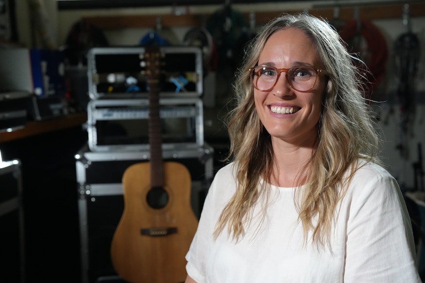 A blonde woman with glasses smiles are the camera, music equipment can be seen in the background.